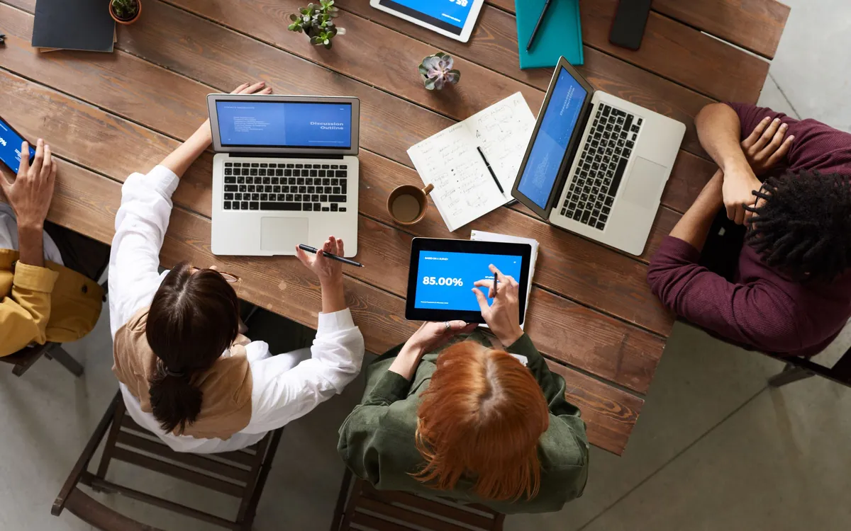 Overhead view of a team meeting around a wooden table with two MacBooks and tablets showing charts