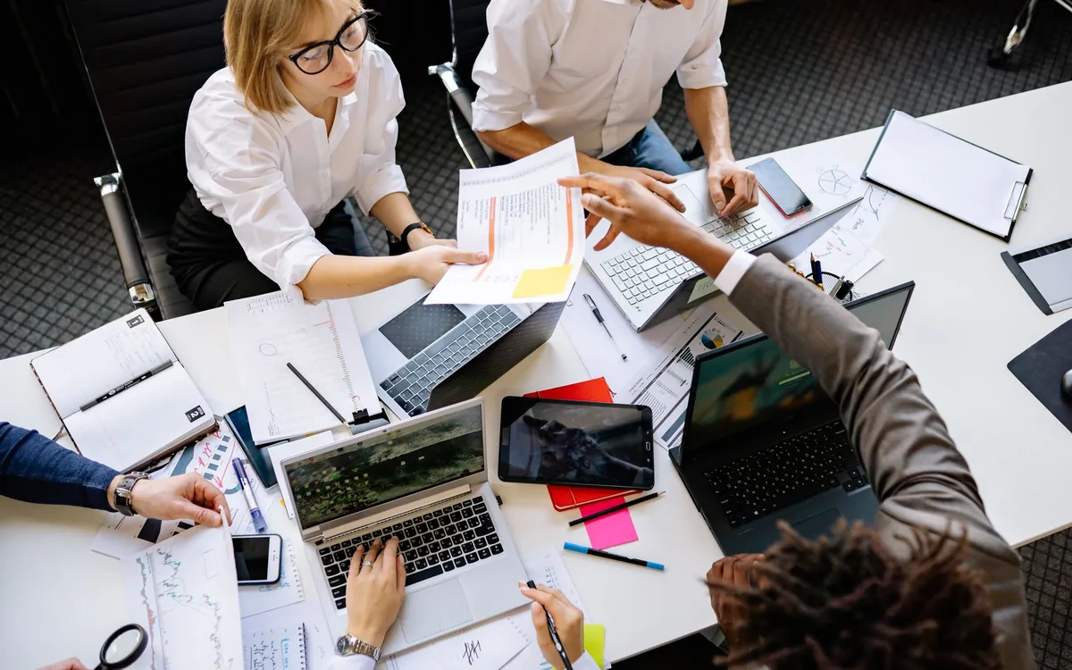 Overhead view of a team collaborating at a shared desk with laptops, a tablet, and printed charts